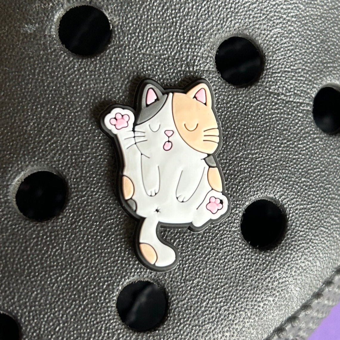 Calico cat licking its bum croc like shoe charm. Shown on a black croc shoe.