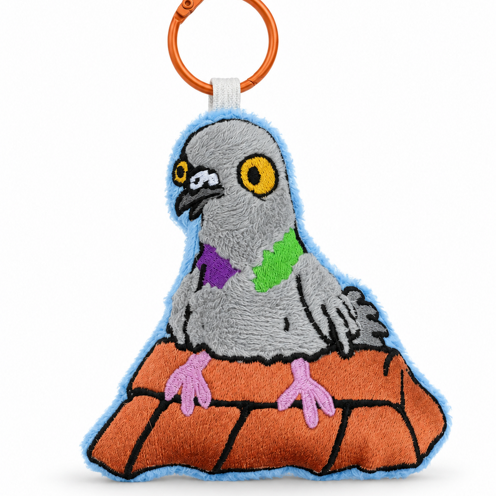 A plush embroidered pigeon keychain or bag charm with a bright orange metal split ring. The pigeon is gray with yellow eyes, wearing a purple and green bow tie, and standing on an orange brick-like mound outlined in black.