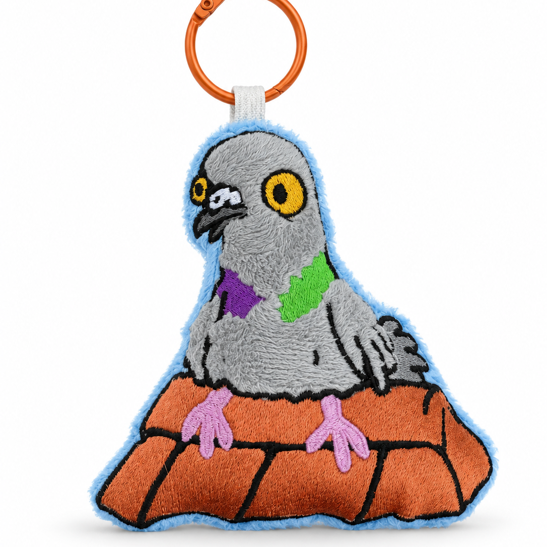 A plush embroidered pigeon keychain or bag charm with a bright orange metal split ring. The pigeon is gray with yellow eyes, wearing a purple and green bow tie, and standing on an orange brick-like mound outlined in black.