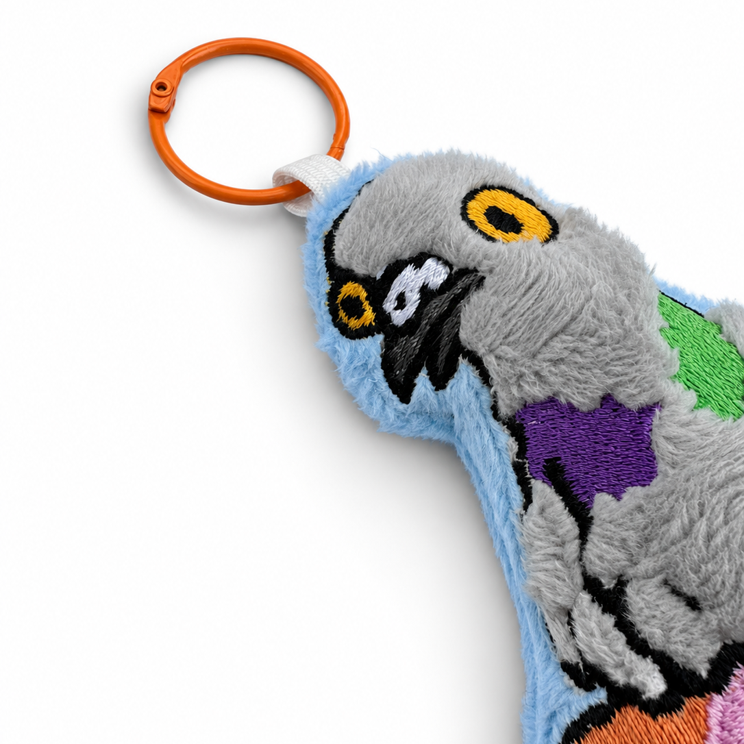 A close-up of a plush toy or bag charm featuring a stylized gray bird character with embroidered yellow eyes, black beak details, and colorful patches in purple, green, orange, pink, and light blue. An orange metal key ring is attached at the top, shown on a clean white background.