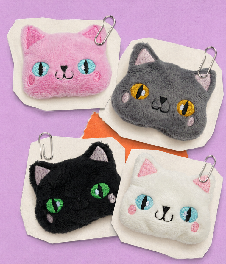 Four small plush cat-face fidgets arranged on a lavender background, each attached to a white paper backing with a metal paperclip. The cats are pink, gray, black, and white, with embroidered eyes and facial features.