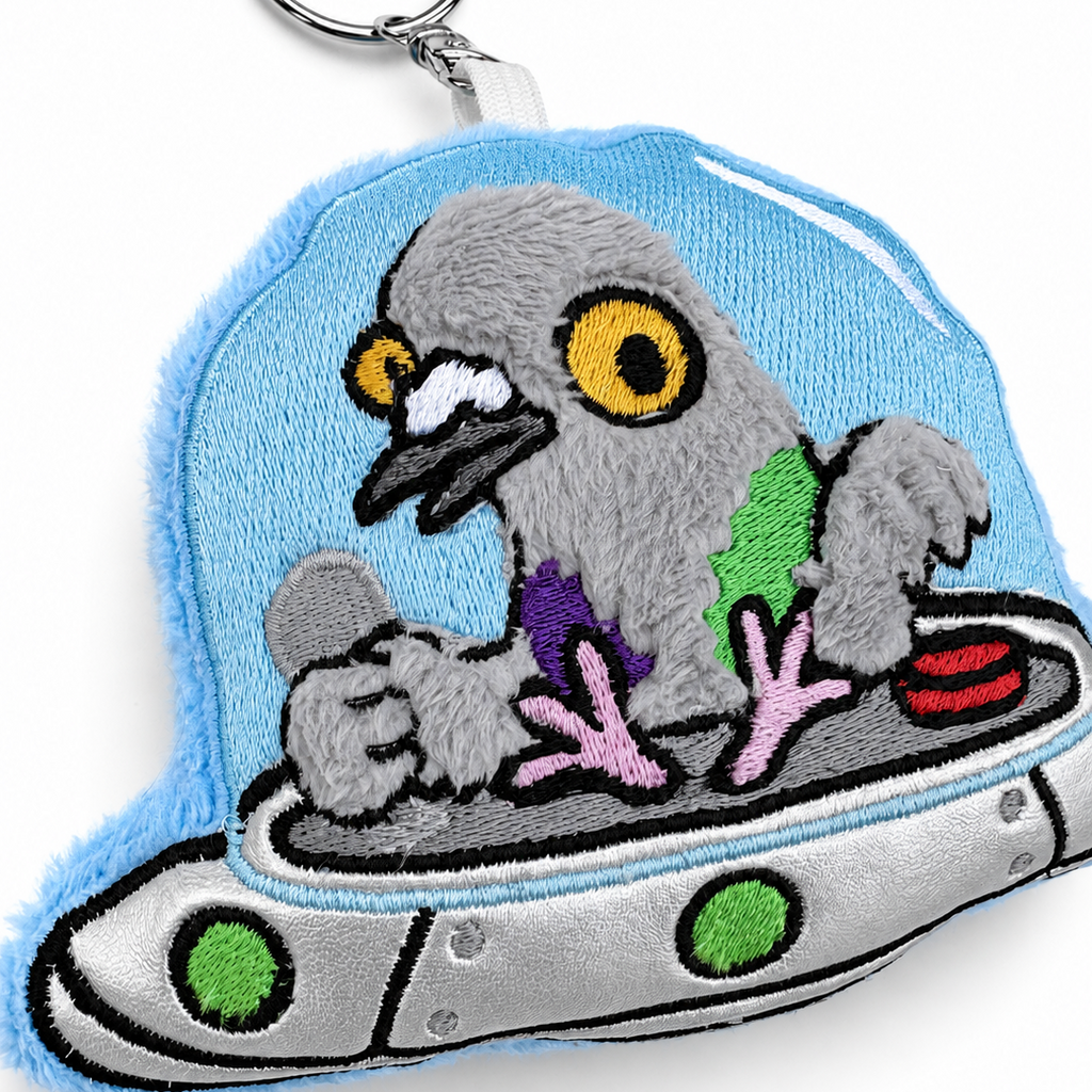 Embroidered plush keychain featuring a gray pigeon in a silver spaceship with a light blue fuzzy outer layer, shown on a white background.