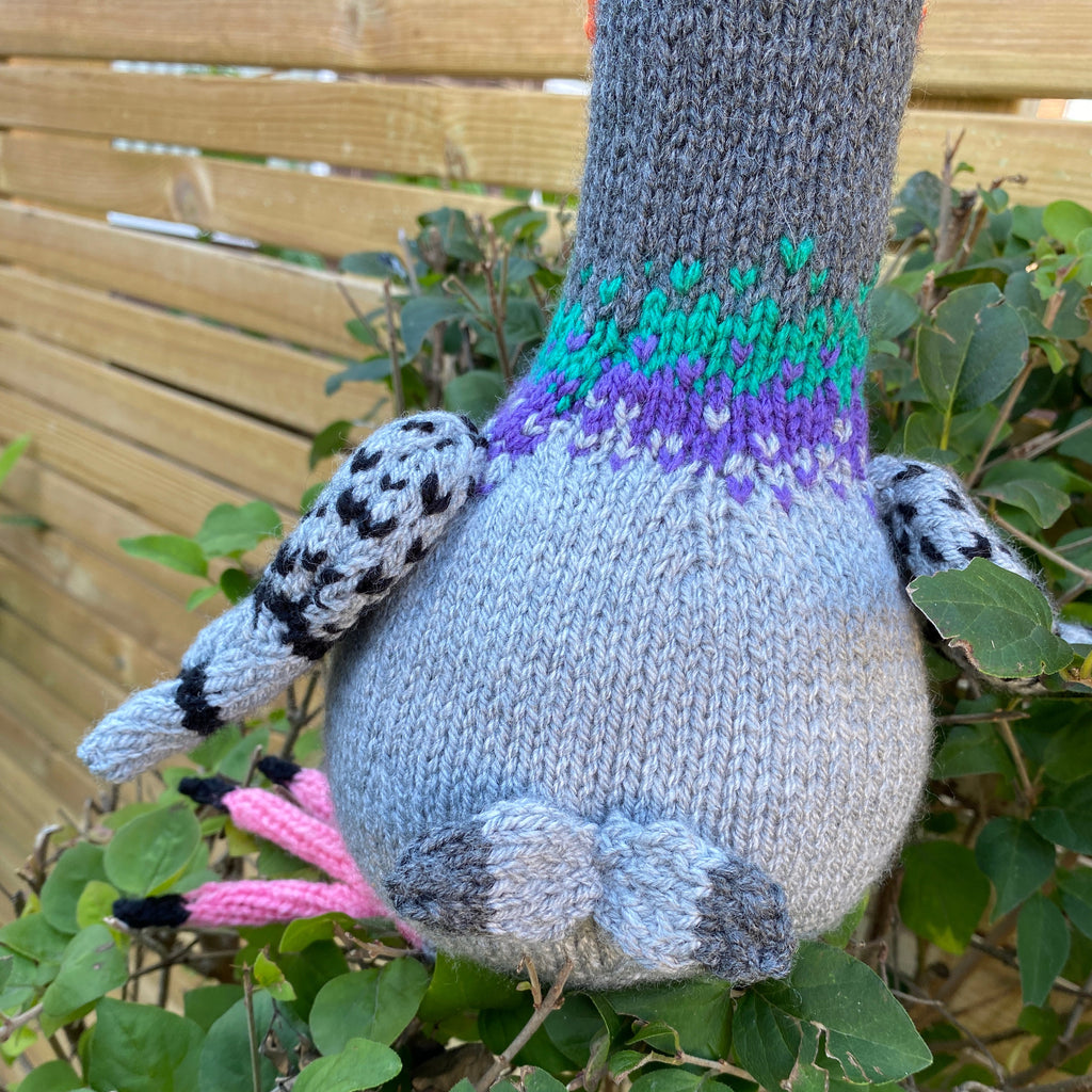 Knitted pigeon toy hanging from a branch with green leaves showing back and tail