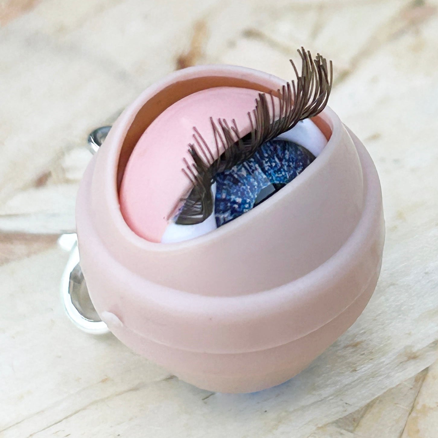 Dolls eye stitch marker shown half closed on a wooden background. 