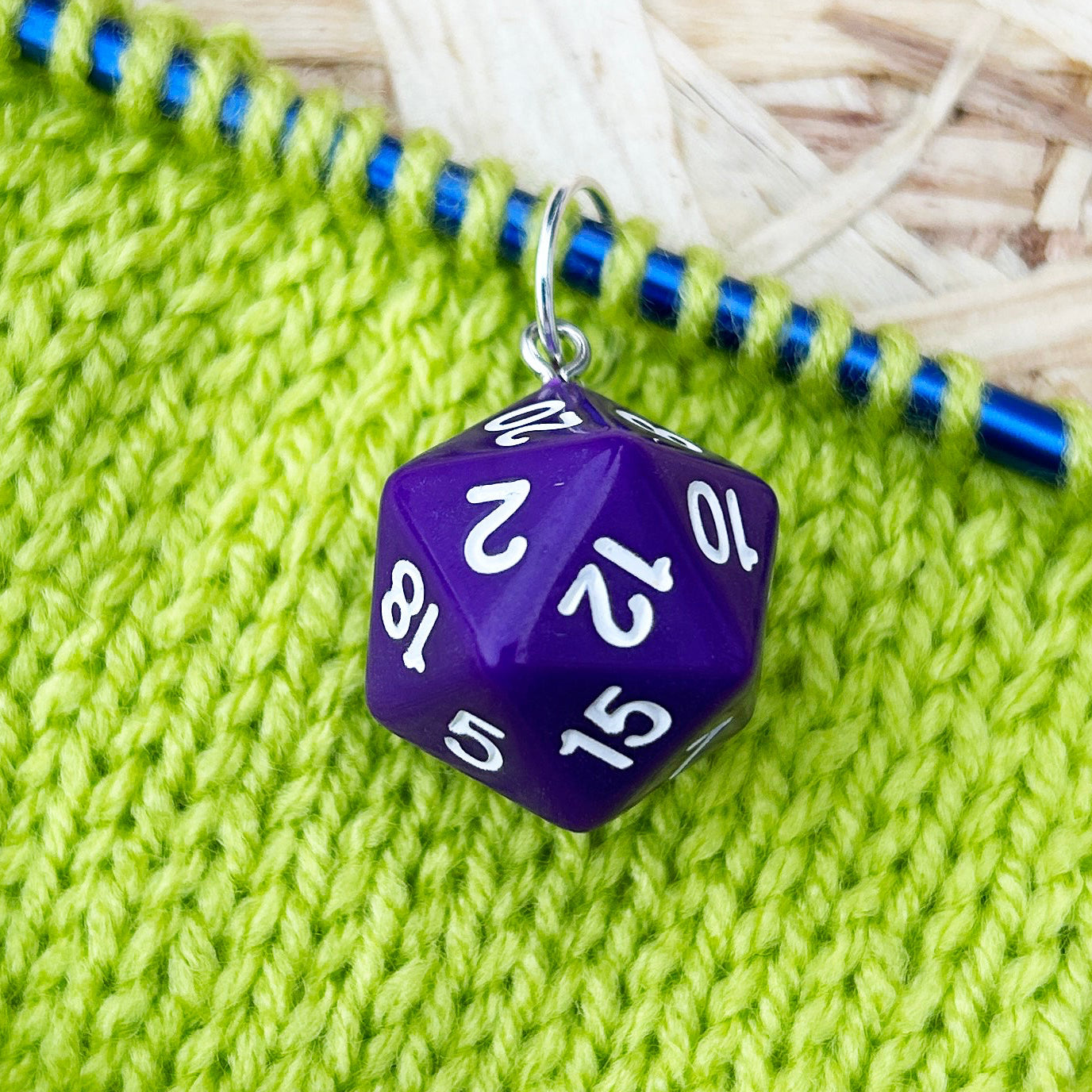 Green knitted fabric with a blue knitting needle and purple dice stitch marker on a straw background