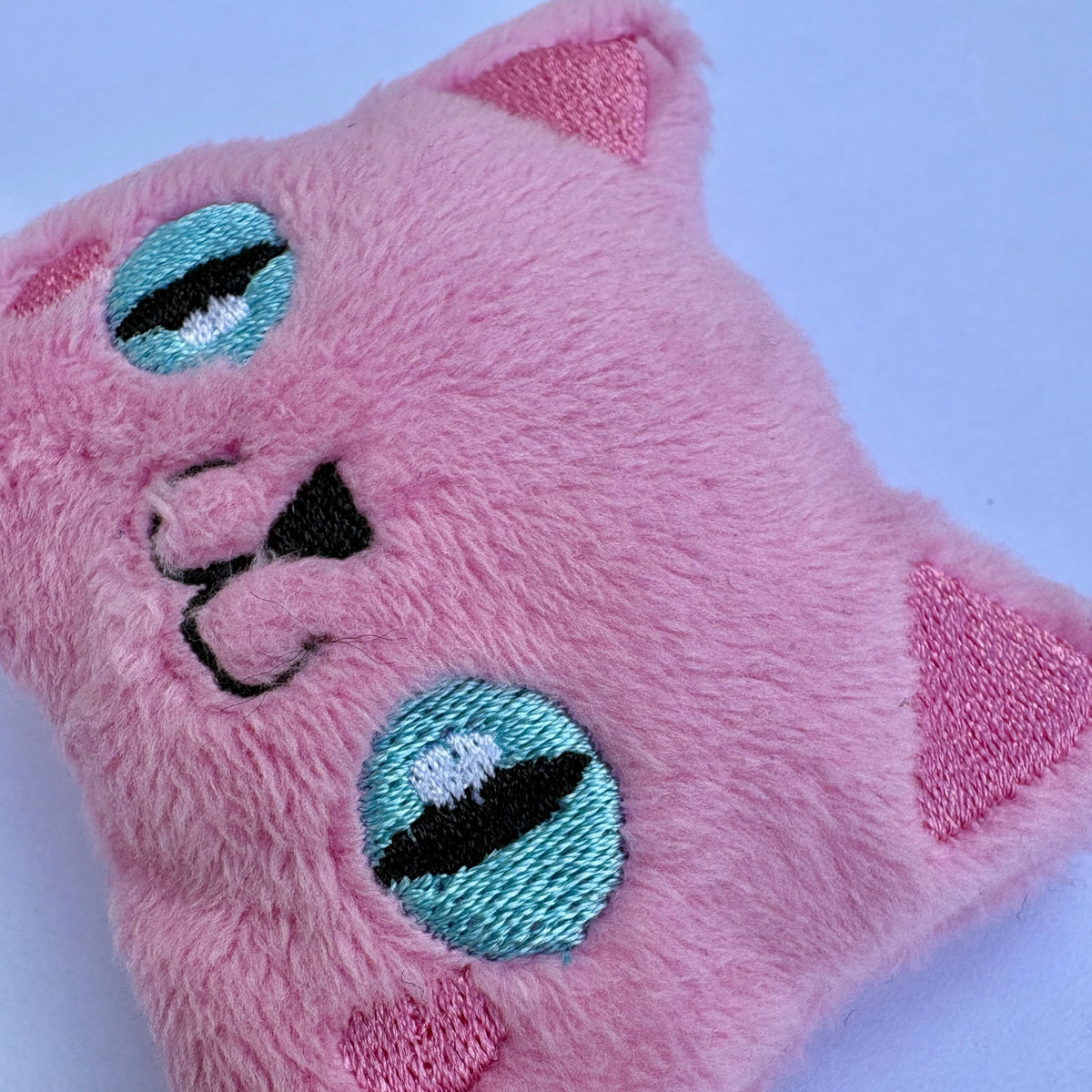 A pink plush cat-shaped cushion or pillow with embroidered facial features, including large turquoise and black eyes, pink ears, and pink paws, shown on a light blue background.