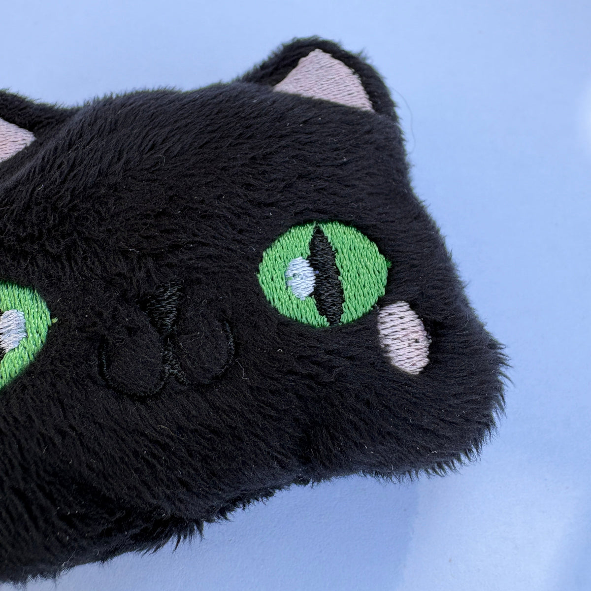 Close-up of a black plush cat toy with embroidered green eyes, pink inner ears, and pink cheek details on a light blue background.
