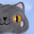 Close-up of a gray plush cat face with embroidered yellow eyes, pink inner ears, and pink cheek spots against a light blue background.