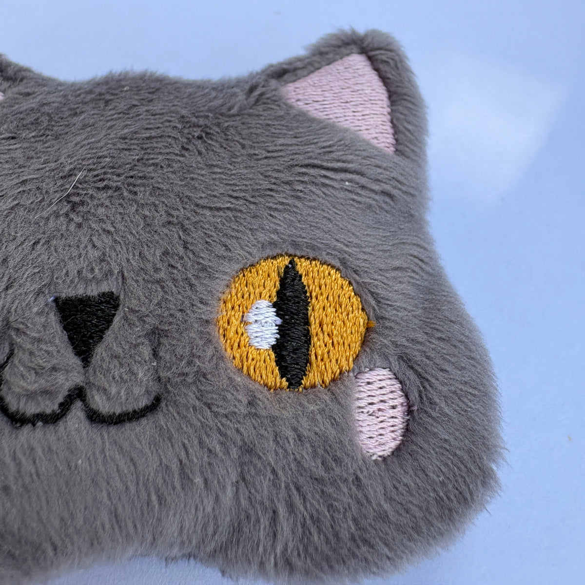 Close-up of a gray plush cat face with embroidered yellow eyes, pink inner ears, and pink cheek spots against a light blue background.