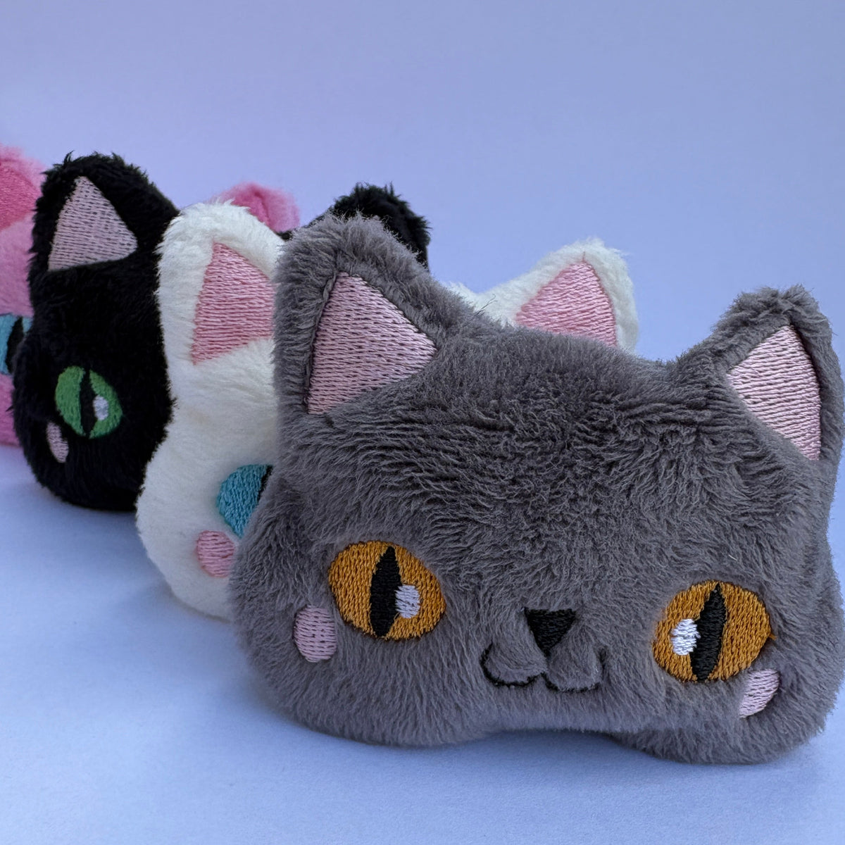 Four plush cat-shaped accessories lined up on a light blue surface, including gray, black, white, pink, and dark blue options, each with embroidered eyes and pink inner ears.