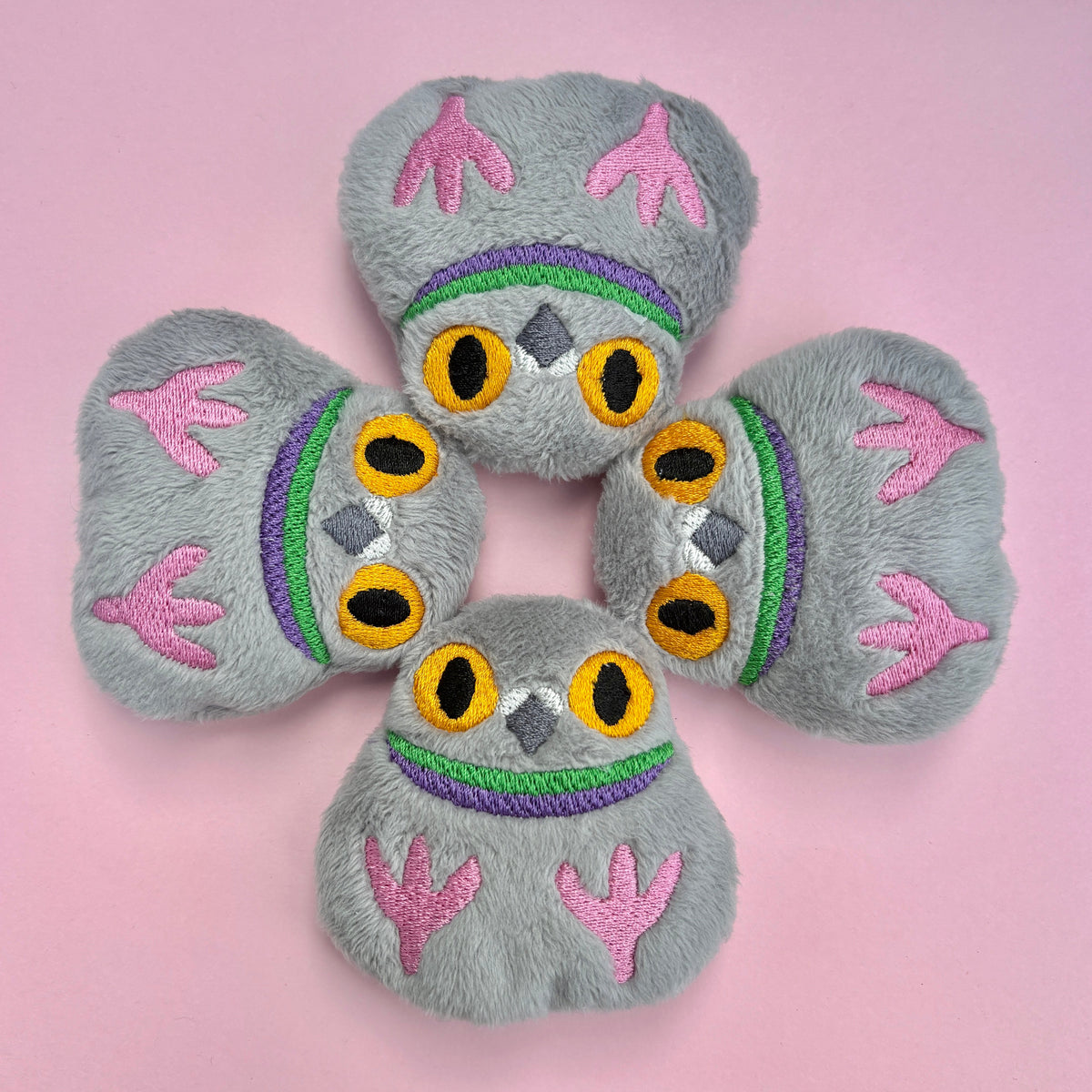 A gray plush pigeon-shaped accessory with embroidered  faces, pink feet, and colorful green and purple stripes, displayed on a pink background.