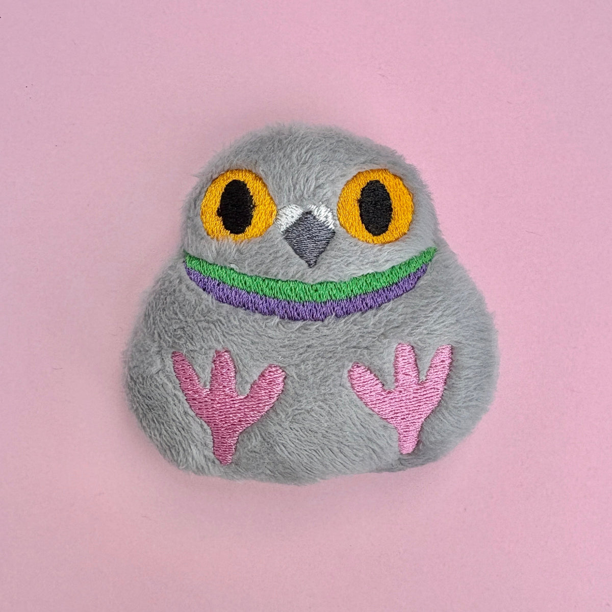 A small gray plush pigeon fidget toy with embroidered orange eyes, a purple and green striped neck, beak, and pink feet, placed on a pink surface.