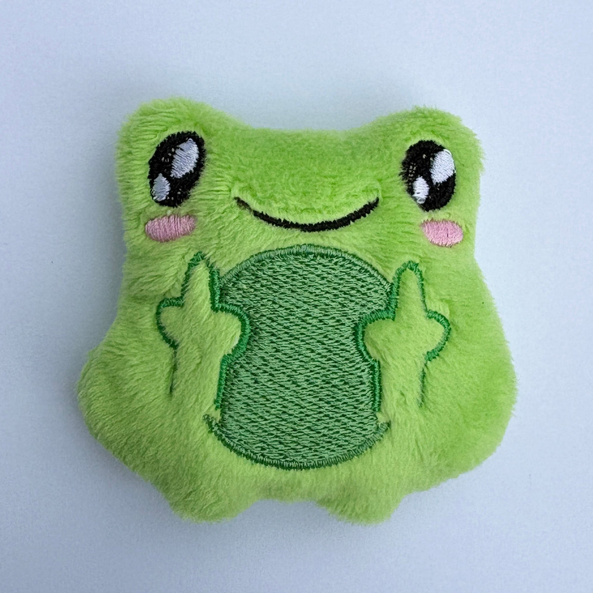 A green plush frog-shaped accessory with embroidered eyes, a smiling mouth, pink cheeks, and raised paws, shown against a plain light background.