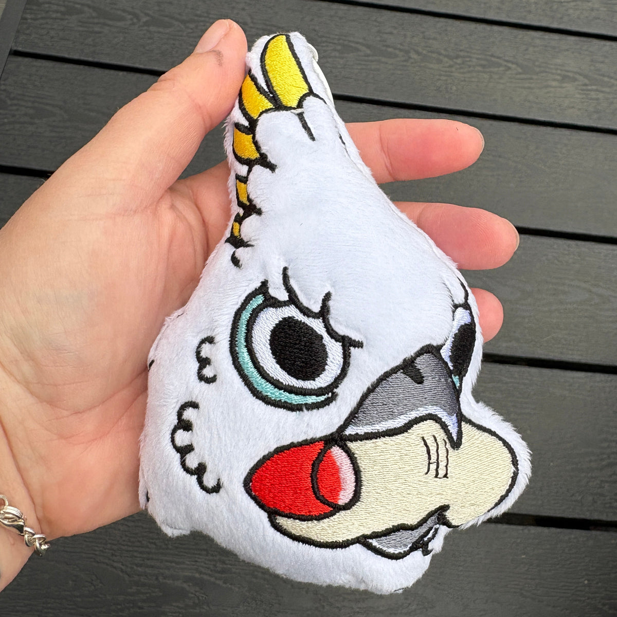 A hand holding a white plush bird bag charm with embroidered facial features, set against a dark gray wooden slatted background.