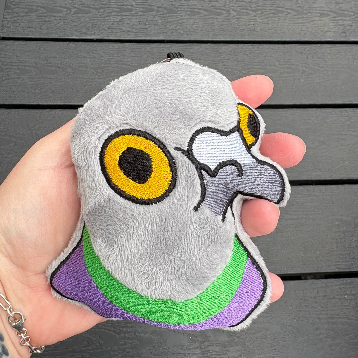 A hand holds a small plush pigeon toy with embroidered gray feathers, a yellow eye, white beak, and purple and green neck details, photographed over a dark gray slatted table.