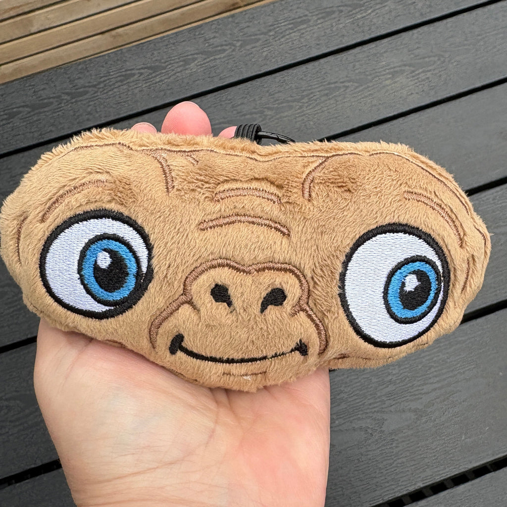 A hand holding a plushET alien shaped bag charm featuring large blue eyes and a brown textured face, set against a dark wooden slatted background.