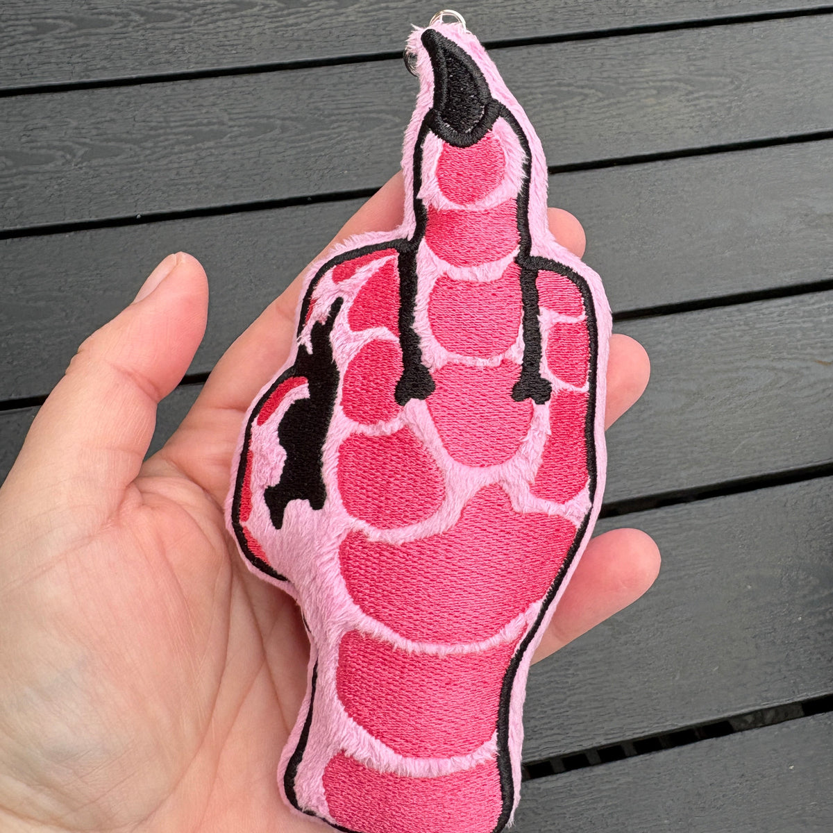 A hand holding a pink and black embroidered plush pillow shaped like a hand gesture, set against a dark gray wooden slat background.