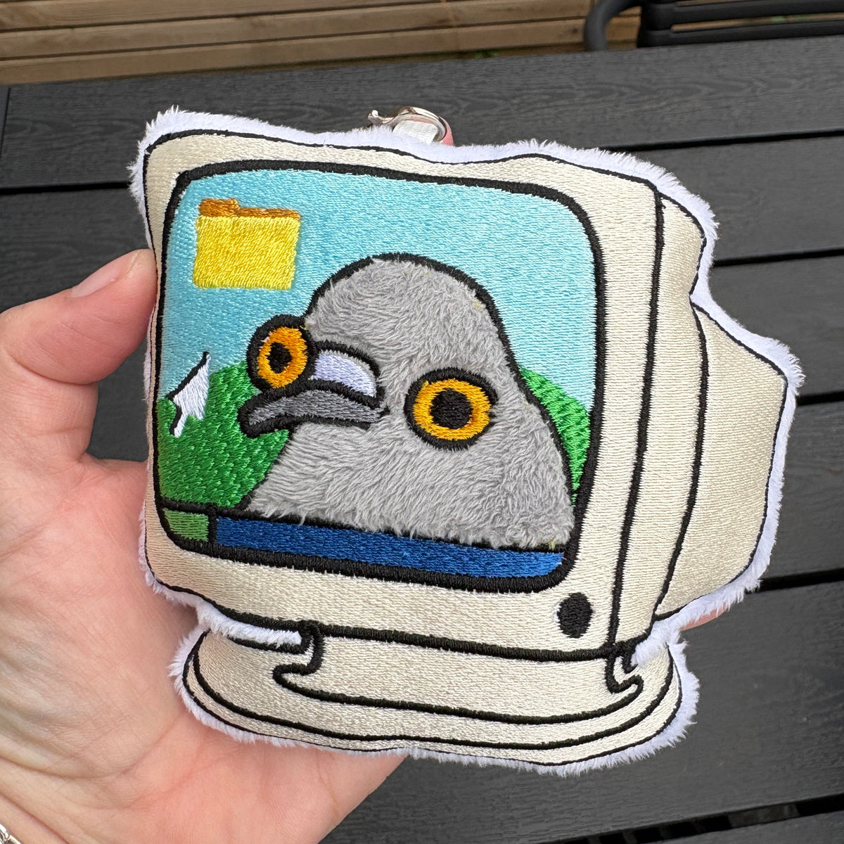 A hand holds an embroidered bag charm/ keychain shaped like a retro computer monitor. The design shows a gray pigeon with large yellow eyes on a light blue screen background, with a cream-colored monitor body and white border.