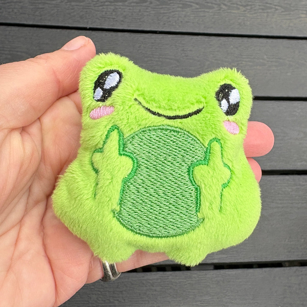A hand holding a small, plush green frog-shaped accessory with embroidered eyes, a smile, and pink cheeks, set against a dark gray wooden slatted background.