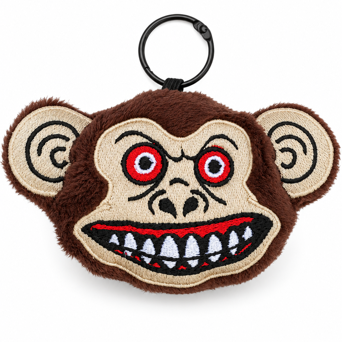 A plush monkey head keychain or bag charm with a black metal key ring. The design features brown fuzzy fur, beige embroidered skin, red and black embroidered eyes, and a wide grin showing white and black teeth.