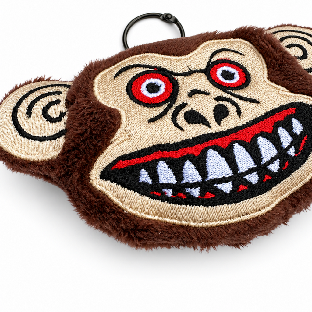 A plush monkey face keychain or bag charm with a black metal ring at the top. The design features brown faux fur, beige embroidered skin, red eyes, and a wide grin showing white and red teeth.