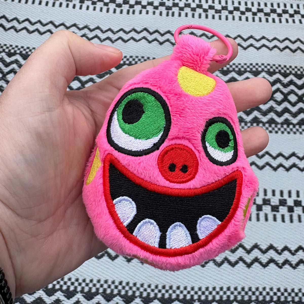 A hand holds a pink plush monster keychain or bag charm with embroidered green eyes, a red nose, yellow accents, and a wide open grin with white teeth, set against a black and white patterned rug.