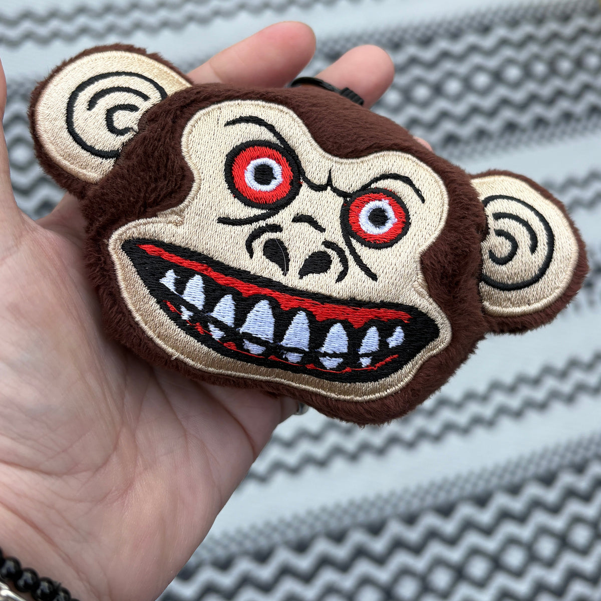 A hand holds a small plush monkey face accessory with a wide grin, red eyes, and spiral ears, photographed outdoors over a patterned rug.