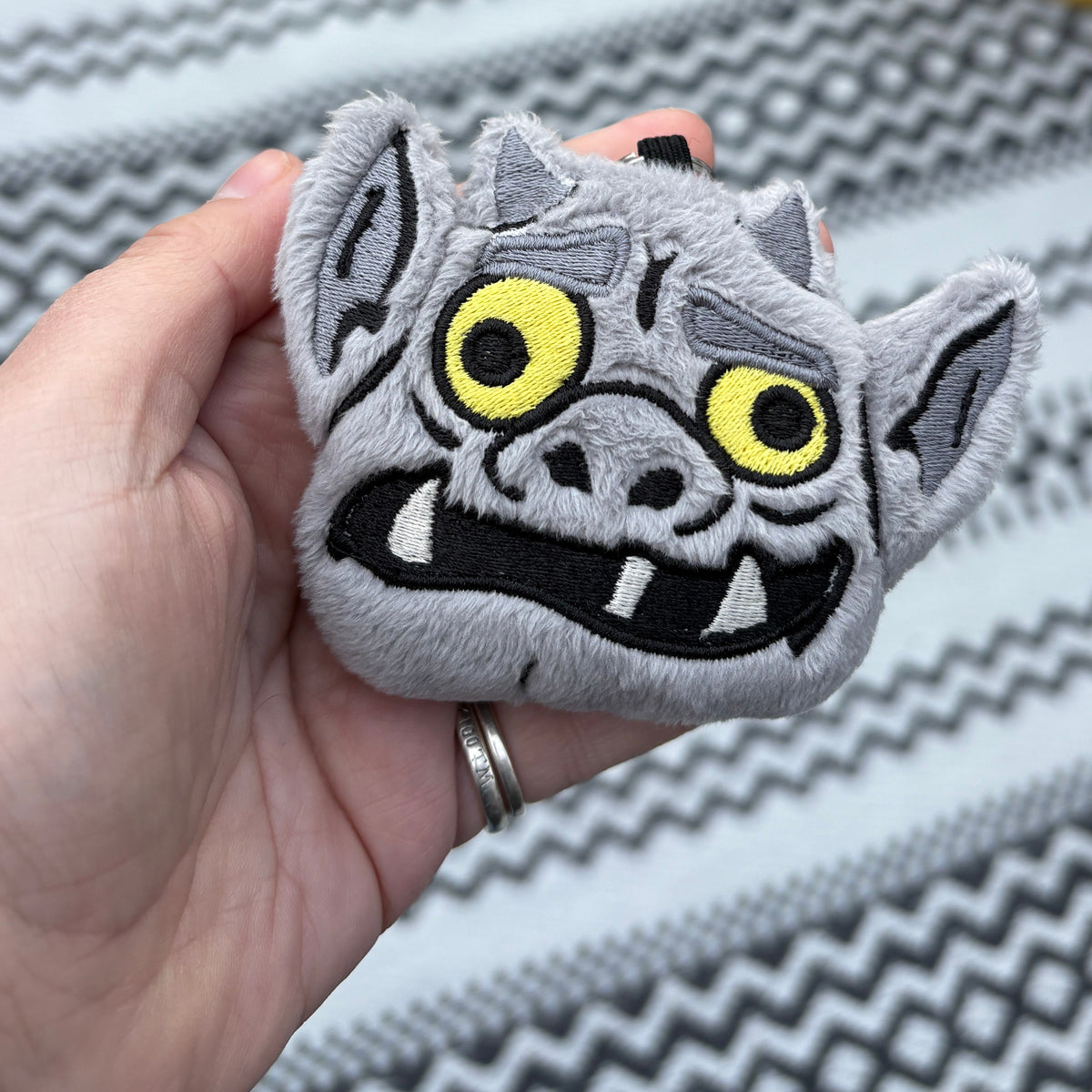 A hand holding a gray embroidered monster face patch with yellow eyes and black stitched details, set against a patterned rug and potted plant in the background.