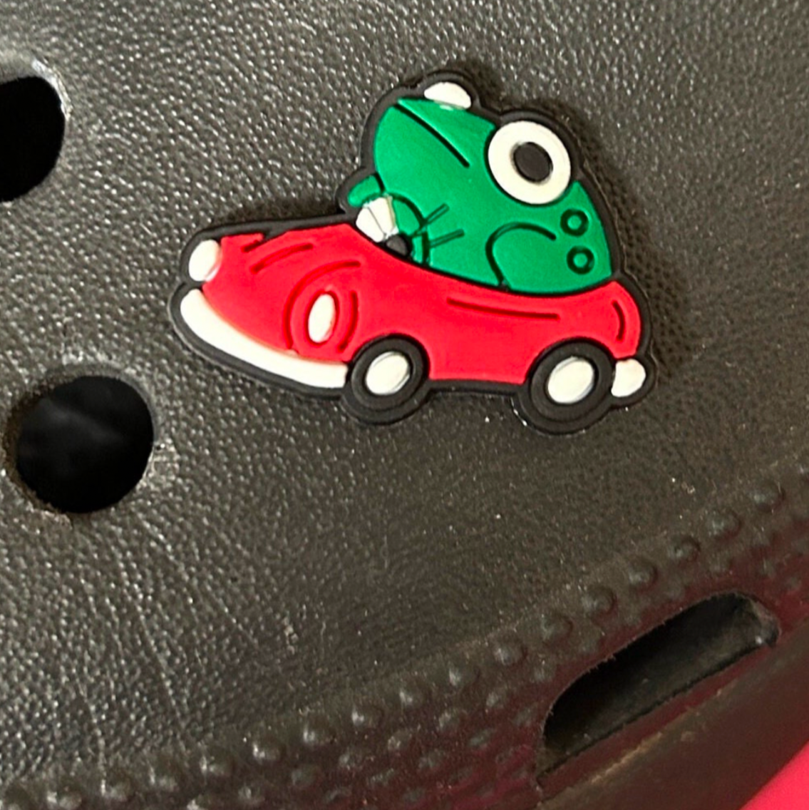 Frog driving a red car croc like shoe charm. Shown on a black croc shoe. 