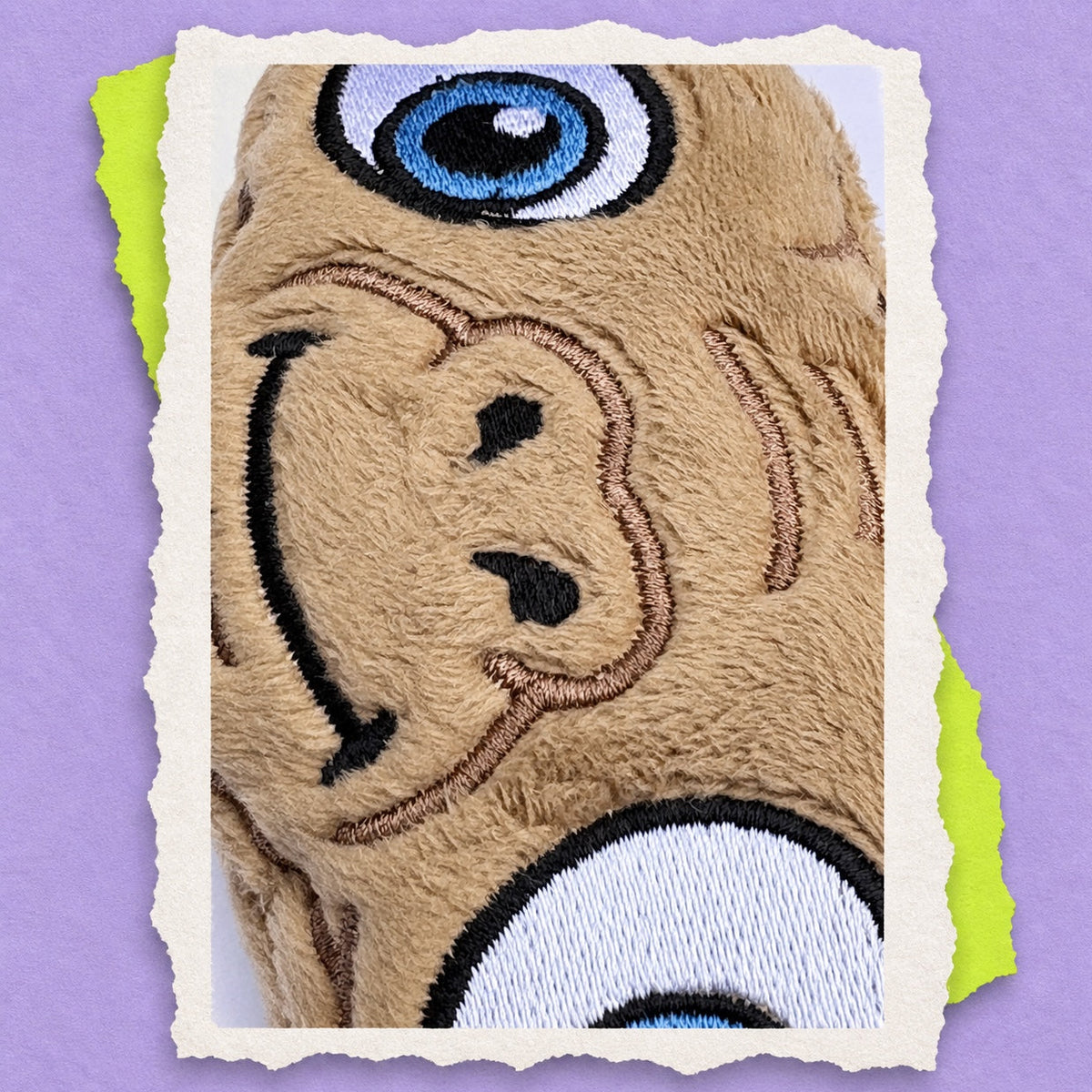 A close-up of a beige embroidered ET plush toy face with a blue eye, black stitched features, and torn-paper style white borders around the photo on a lavender background.