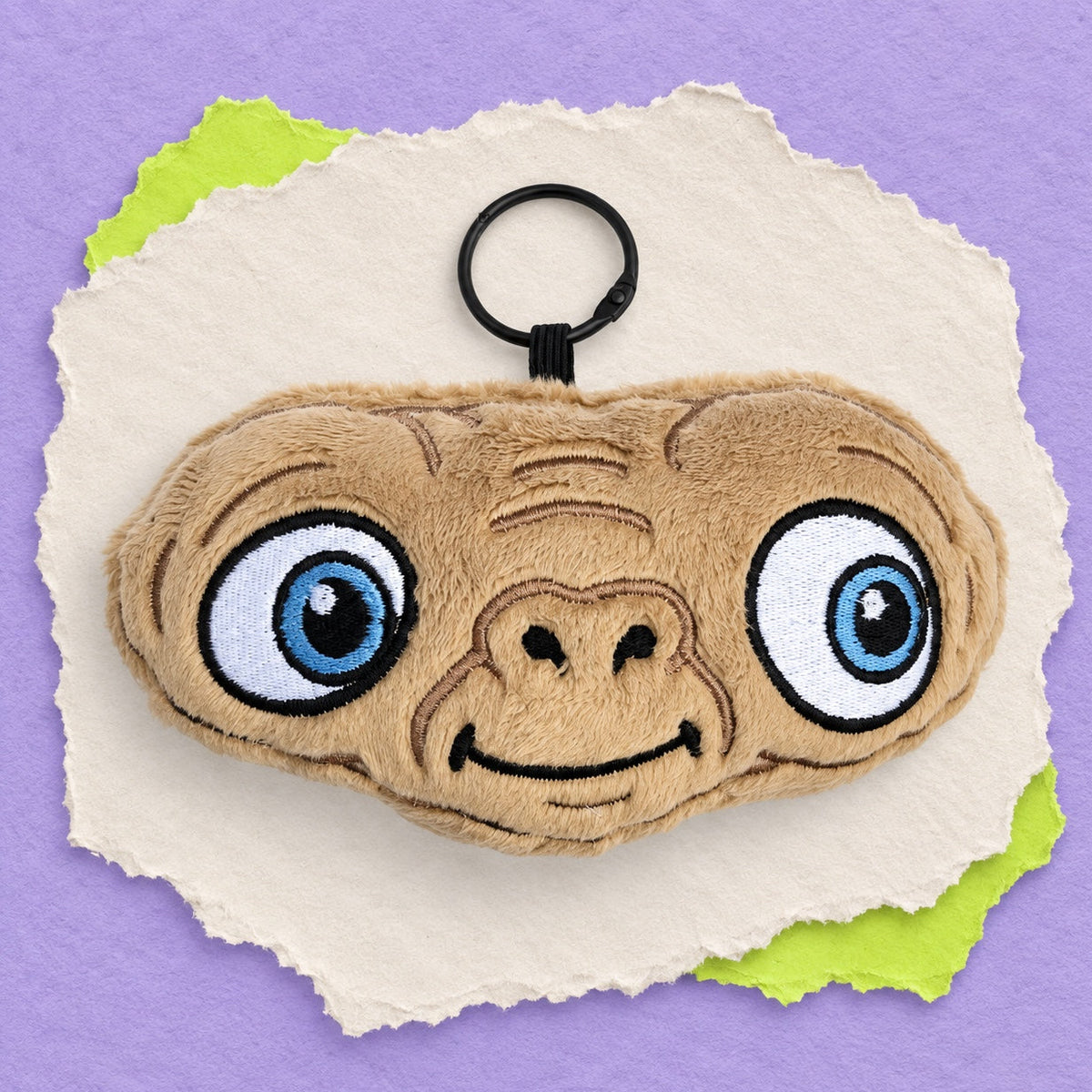 A plush character alien ET face keychain featuring a tan embroidered face with large blue eyes and a black key ring, centered on a torn white paper background with green paper edges against a purple backdrop.