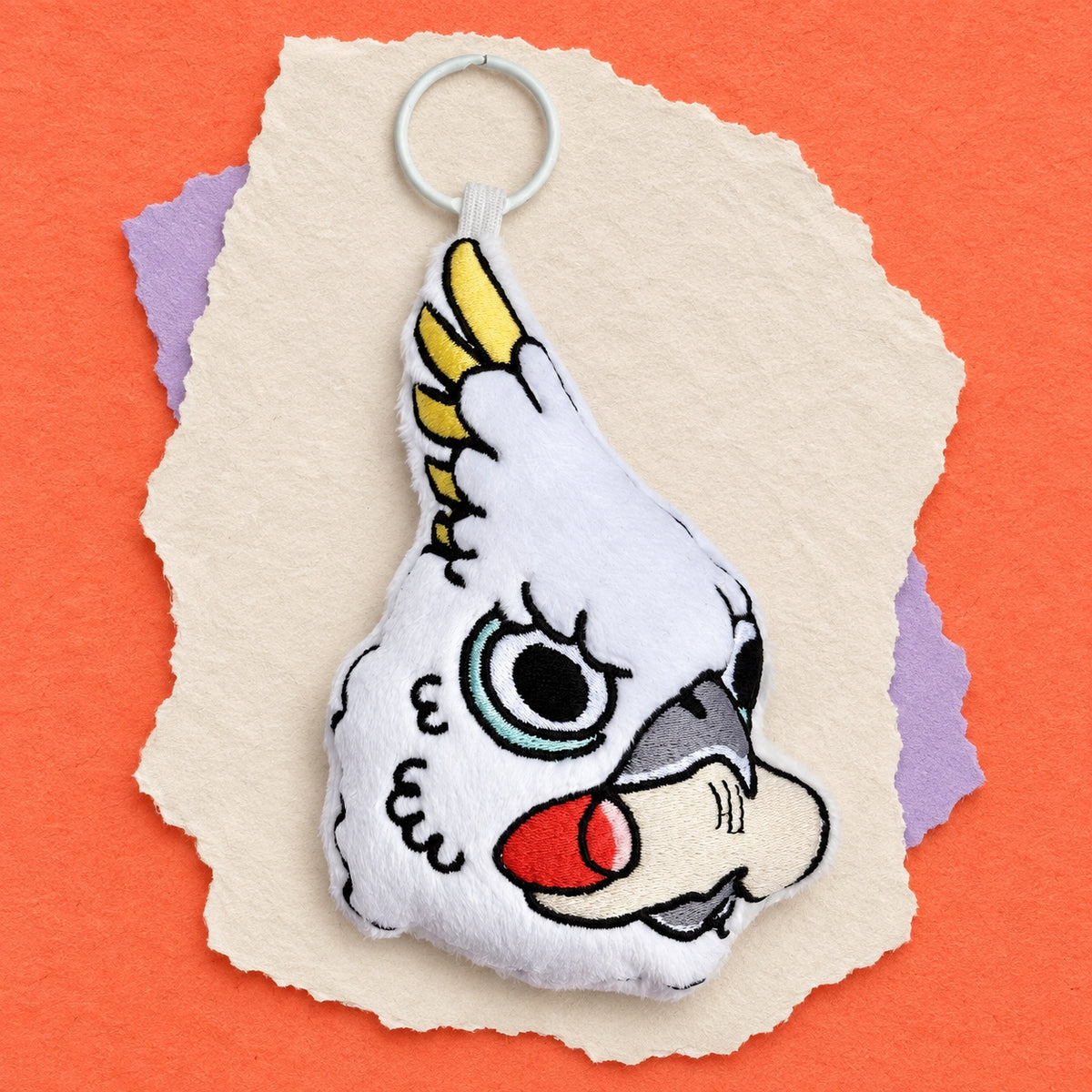 A white embroidered bird plush keychain or bag charm with a metal key ring, featuring a stylized face with yellow, red, gray, and blue details, presented on torn paper over an orange background.