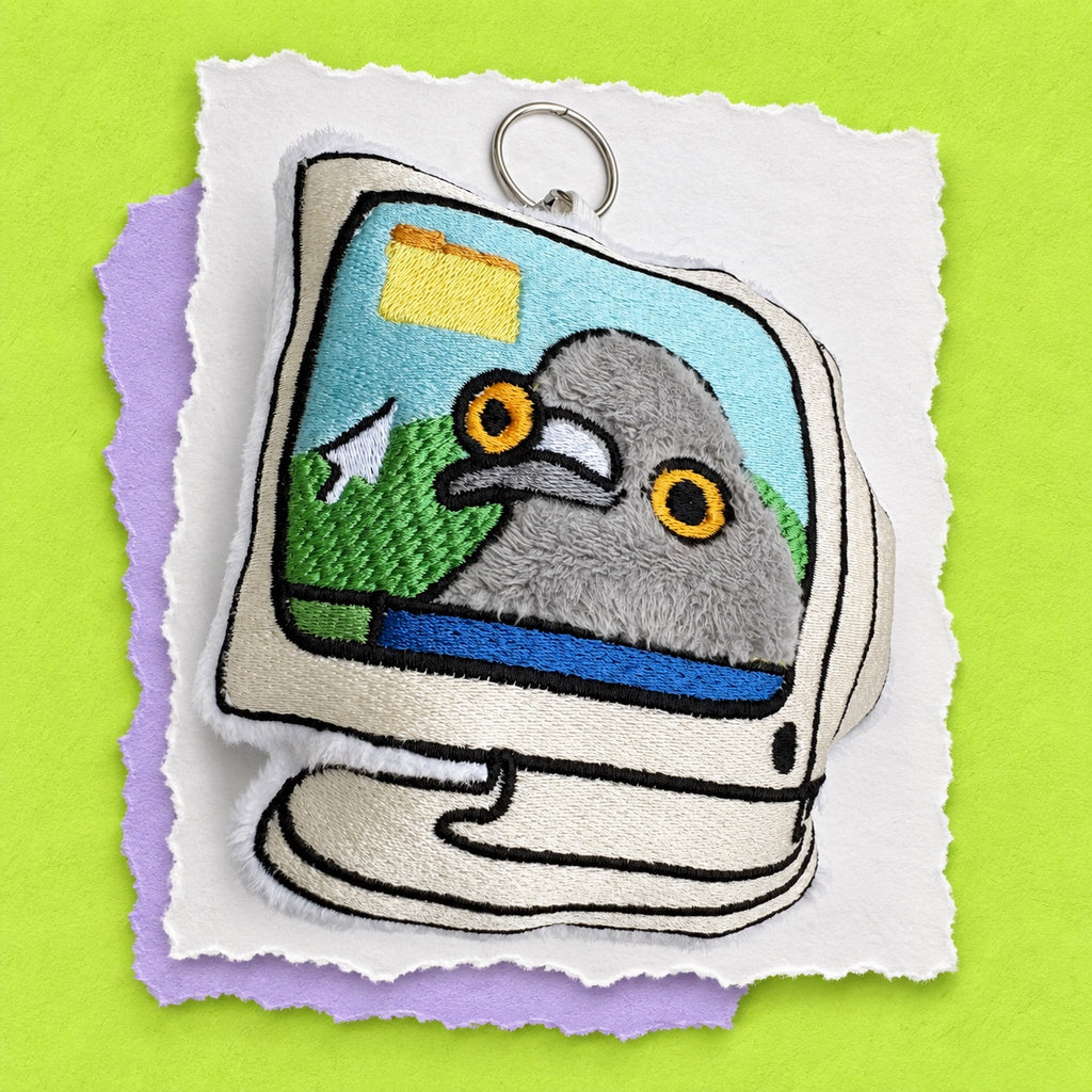 A novelty embroidered plush keychain shaped like a retro computer monitor. The screen displays a gray pigeon with yellow eyes, and the plush body is beige with black embroidered outlines.
