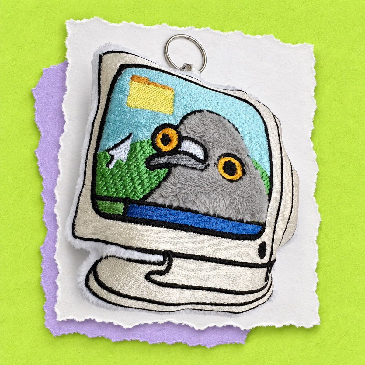 A novelty embroidered plush keychain shaped like a retro computer monitor. The screen displays a gray pigeon with yellow eyes, and the plush body is beige with black embroidered outlines.