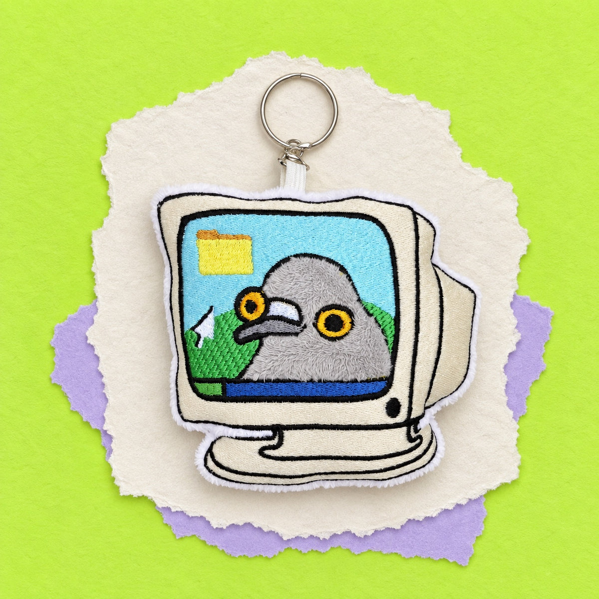 An embroidered keychain shaped like a retro computer monitor with a pigeon character inside the screen, shown on torn paper against a bright lime green background.