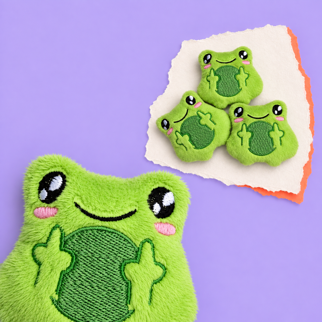 A green plush frog-shaped fidget with embroidered eyes, pink cheeks, and a stitched belly, shown on a lavender background. A torn white paper corner in the upper right holds three smaller matching frog pieces.