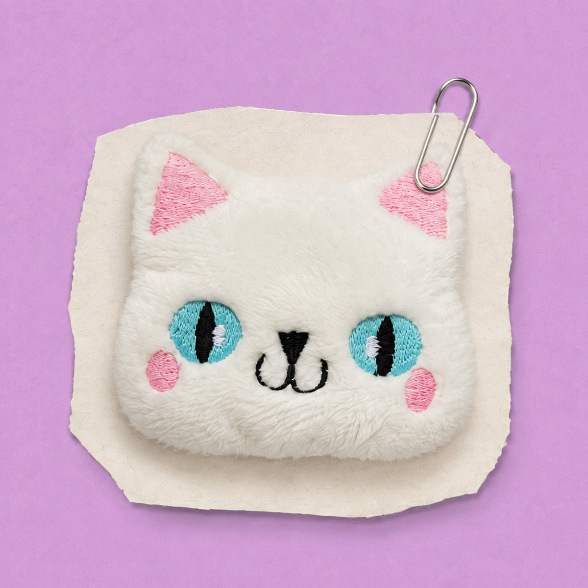 A small white plush cat-face fidget with embroidered blue eyes, pink ear tips, pink cheeks, and a black stitched nose and mouth. A silver paperclip is clipped to the top, and the item is photographed on a lavender background.