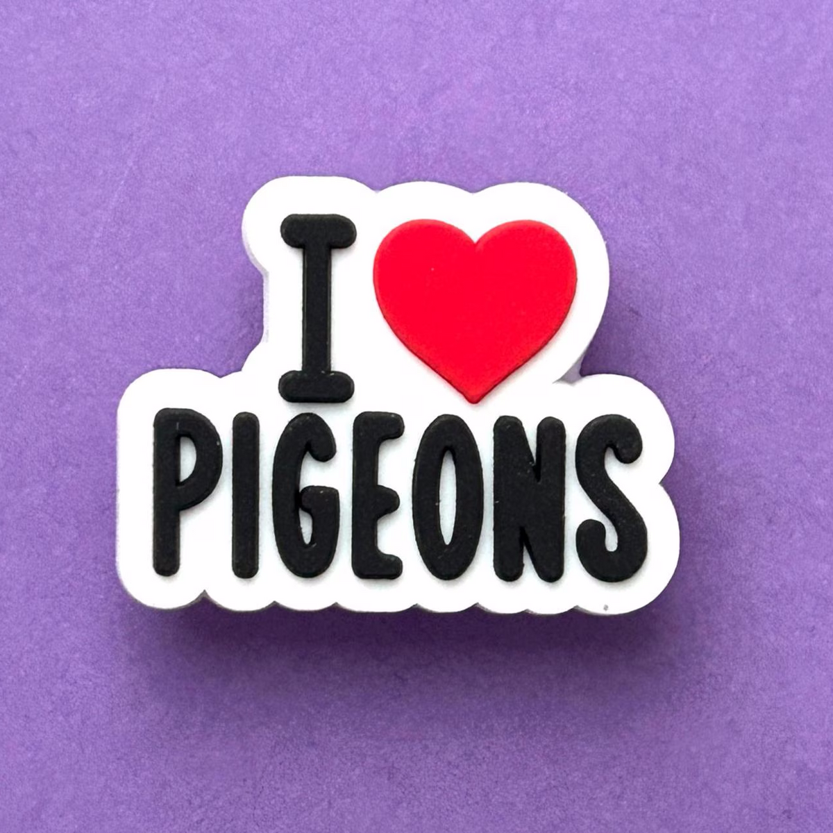 A die-cut enamel pin or sticker with the text 'I' in black, a red heart, and 'PIGEONS' in black, outlined in white, set against a solid purple background.