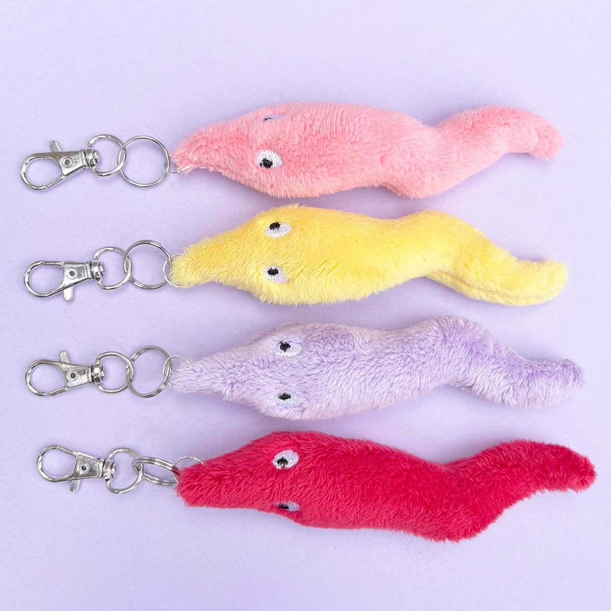 Four plush worm-shaped keychains in pink, yellow, lavender, and red, arranged vertically on a light purple background. Each worm has small embroidered eyes and a silver metal lobster clasp and key ring.