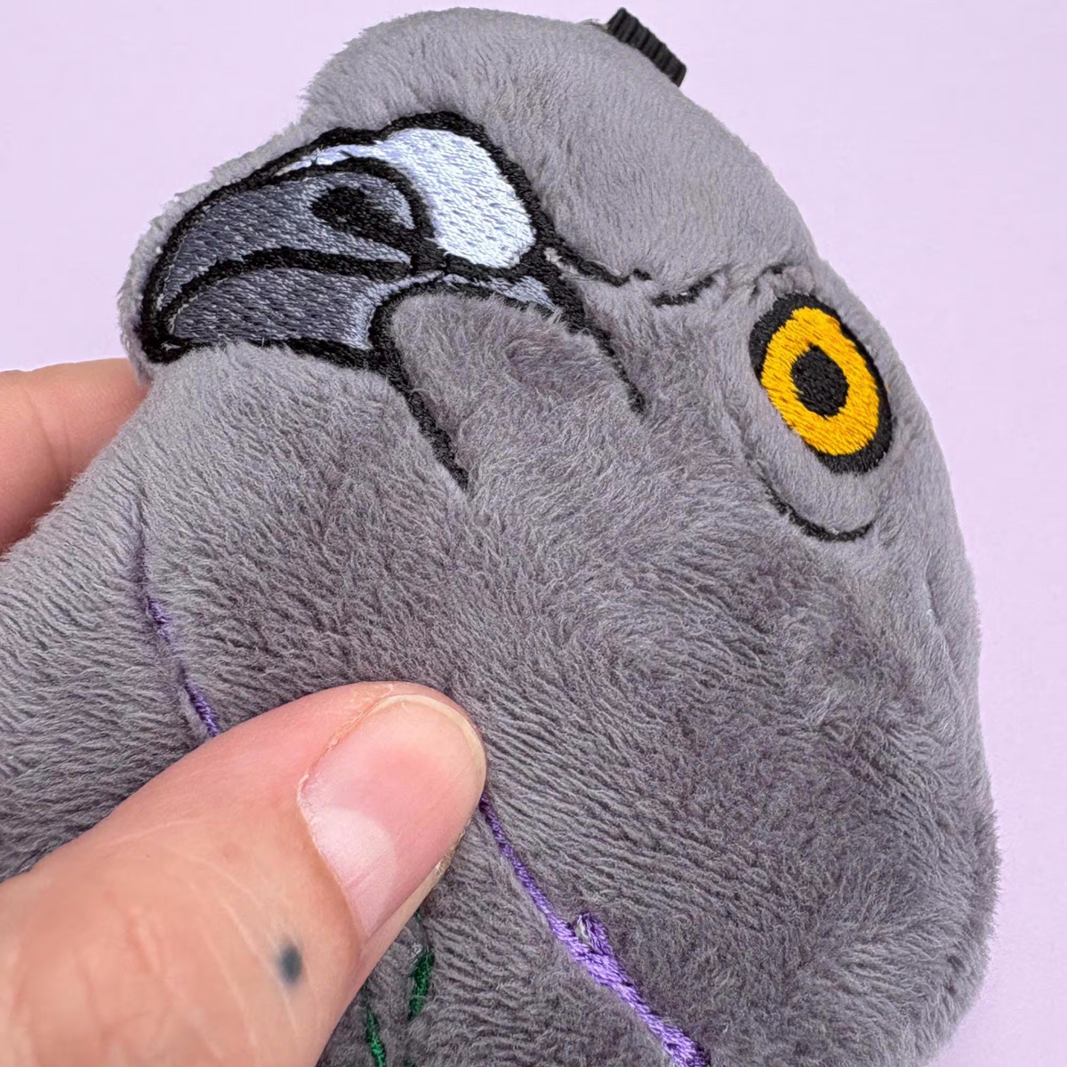 A close-up of a gray plush pigeon toy being held by a human hand, showing embroidered details including a yellow eye, black beak, and purple stitched beak.