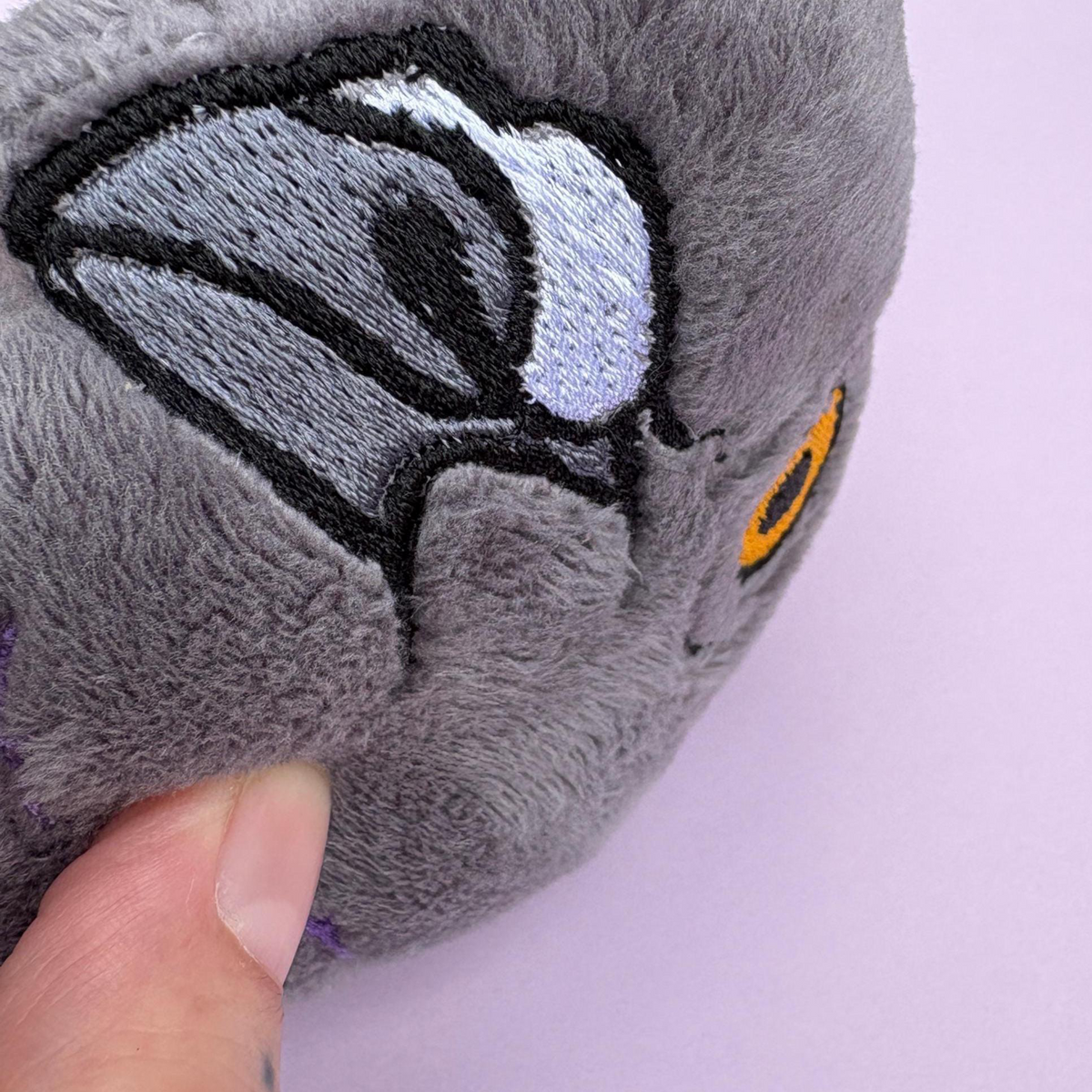 Close-up of a gray plush toy with embroidered details, including a black, white, and gray beak and a small orange eye, held by a person's finger against a light purple background.