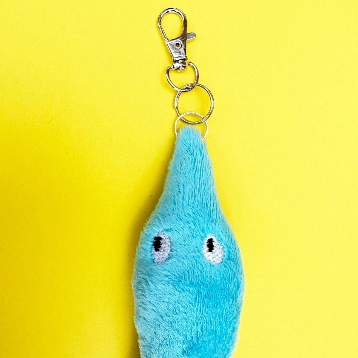 A light blue plush keychain shaped like a teardrop with embroidered eyes, attached to silver metal rings and a lobster clasp, set against a solid yellow background.