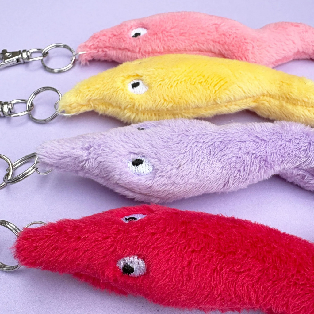 Four plush fish-shaped keychains in pink, yellow, lavender, and red, arranged diagonally on a light purple background.