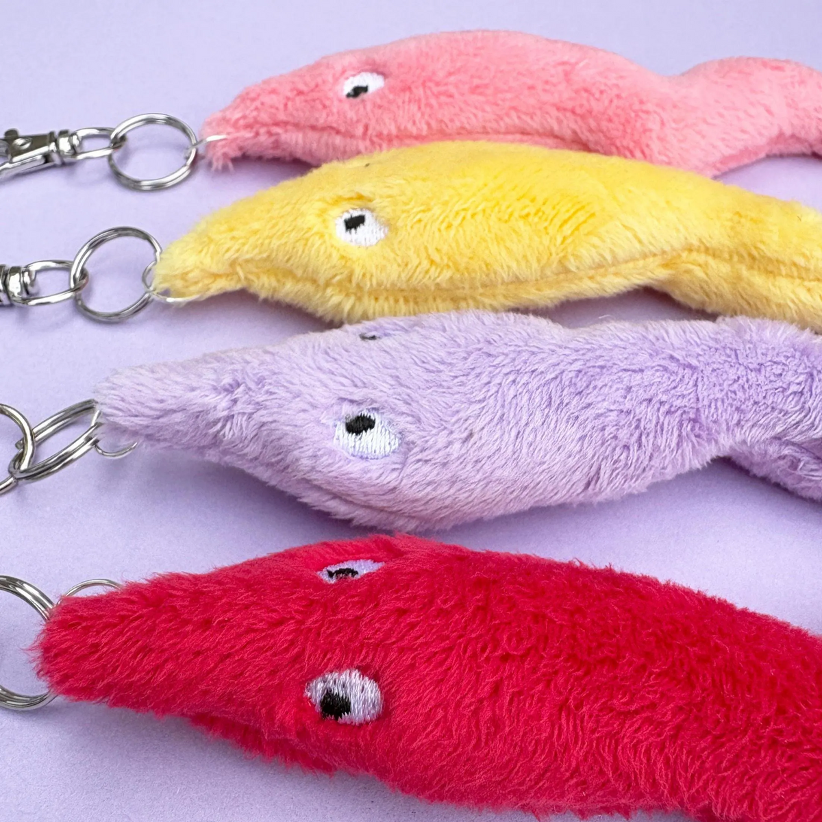 Four plush fish-shaped keychains in pink, yellow, lavender, and red, arranged diagonally on a light purple background.