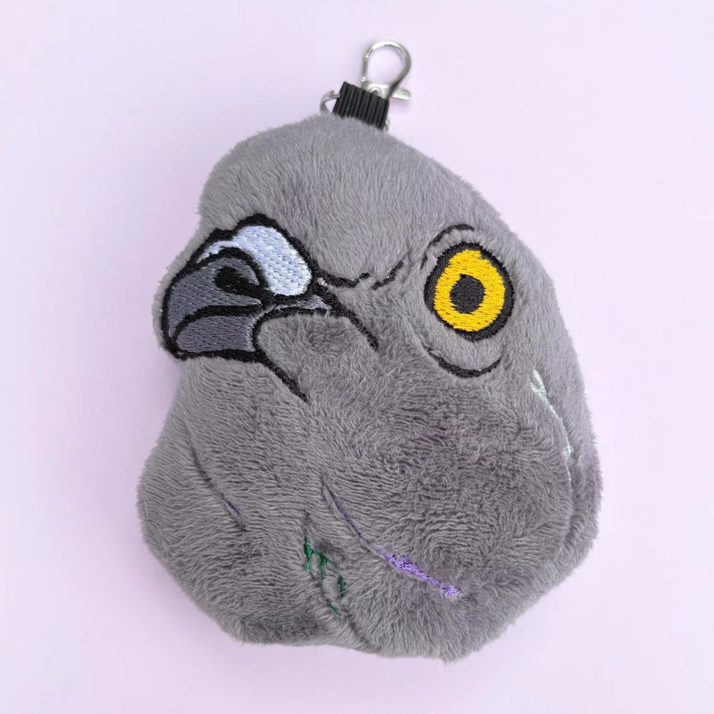 A plush gray pigeon head keychain with embroidered facial details, including a yellow eye, black beak, and silver metal clasp at the top, shown on a pale pink background.