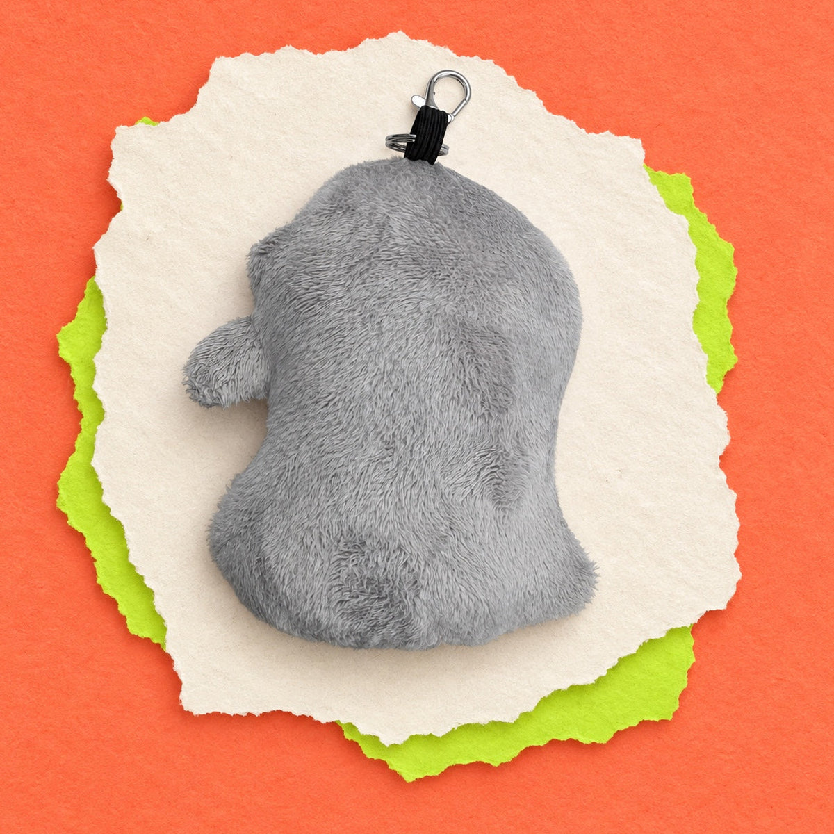 A gray plush keychain back view of pigeon, shown from the back with a black clip, placed on torn white paper over bright green and orange paper layers.