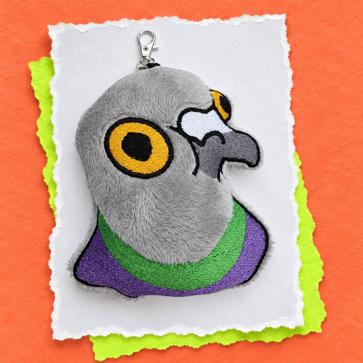 A plush pigeon head keychain or bag charm with embroidered details, shown on a layered white, lime green, and orange paper background.
