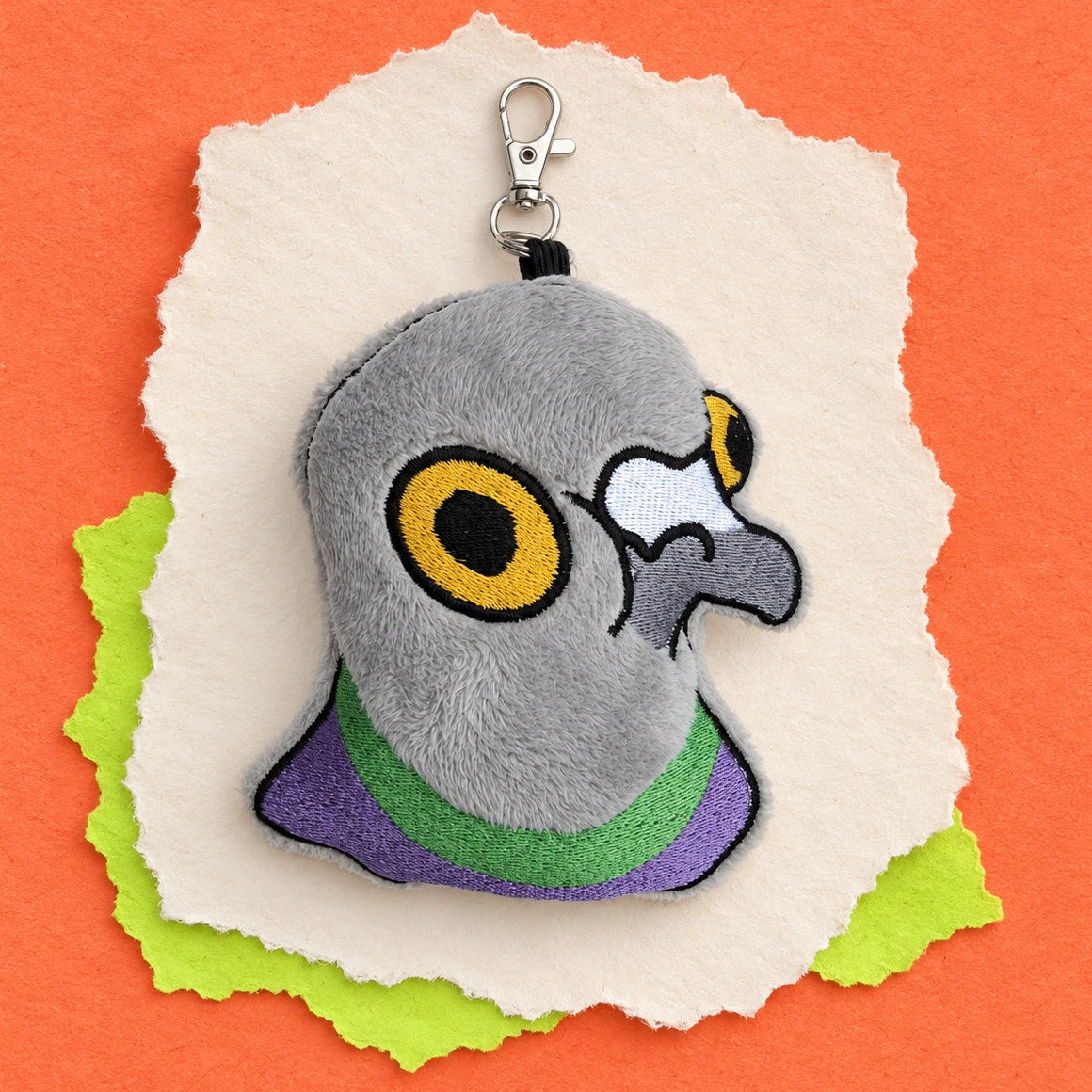 A plush pigeon head keychain or bag charm with embroidered gray feathers, yellow eyes, a purple and green collar, and a silver clasp, displayed on torn white and green paper against an orange background.
