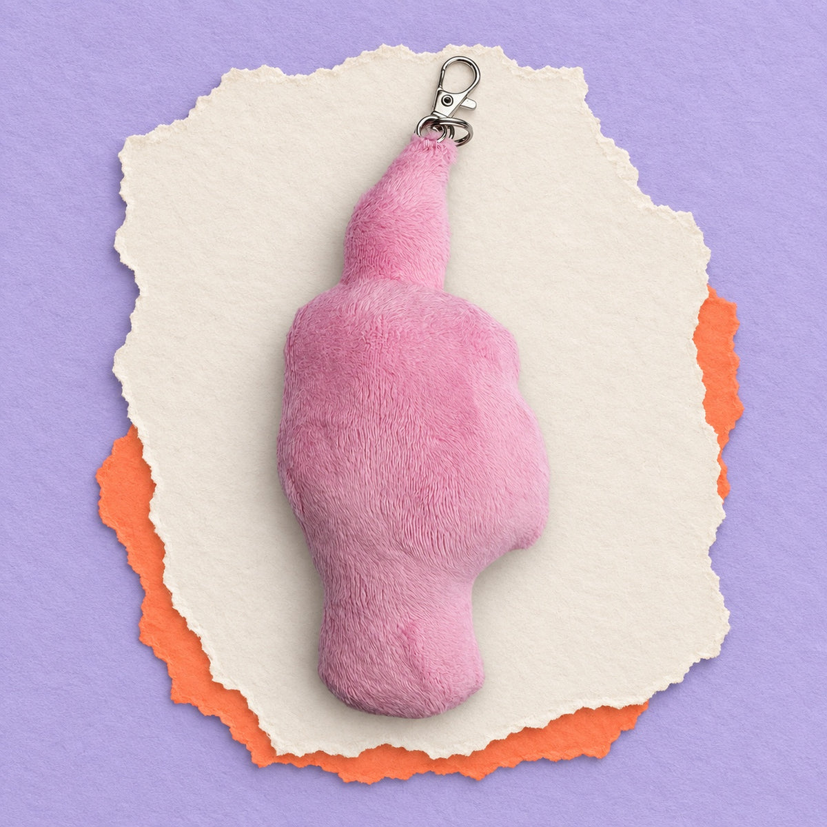 A pink fuzzy novelty keychain shaped like a hand gesture, displayed on torn white paper with an orange paper layer underneath, set against a solid lavender background.