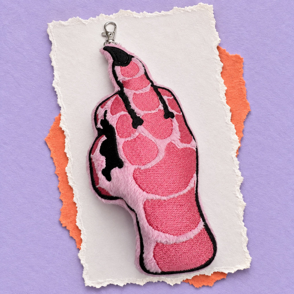 An embroidered keychain charm shaped like a hand giving the middle finger, featuring a pink and black design with a silver lobster clasp, displayed on torn white paper over orange and purple textured backgrounds.